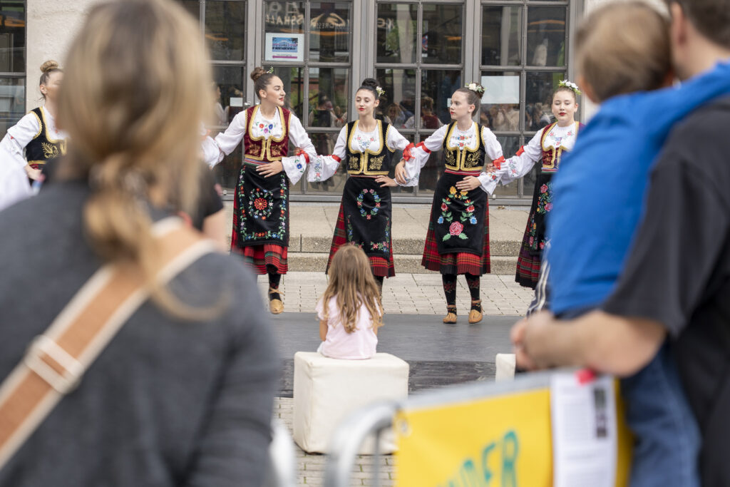 Tänzerinnen aus der südlichen Region Serbiens stehen auf einer Bühne, davor sitzt ein Mädchen auf einem Block und schaut sie an.