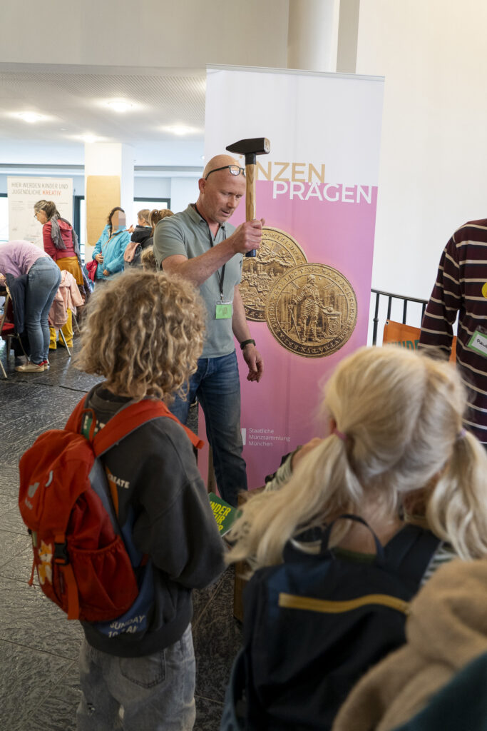 Zwei Erwachsene erzählen einer Gruppe von Kindern etwas, dahinter ein Roll-Up: Münzen prägen.