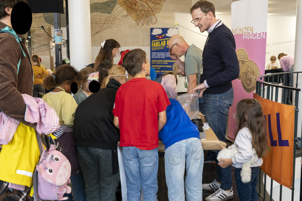 Zwei Erwachsene erzählen einer Gruppe von Kindern etwas, dahinter ein Roll-Up: Münzen prägen.