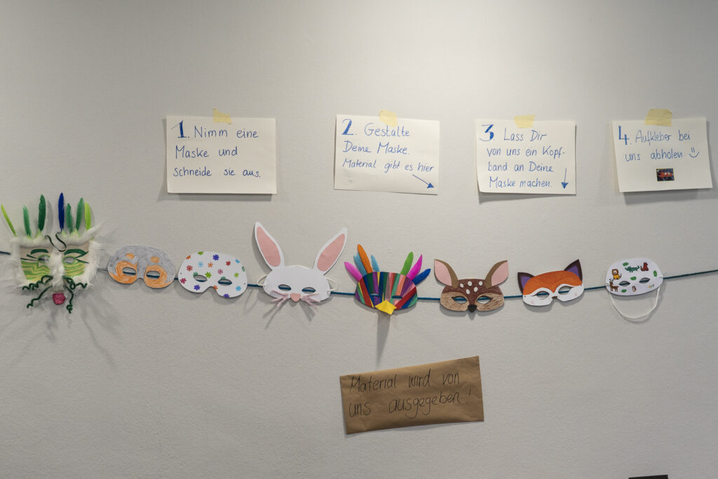 Eine Wand an der verschiedene selbstgemachte Tiermasken hängen und darüber eine kurze Bastelanweisung.
