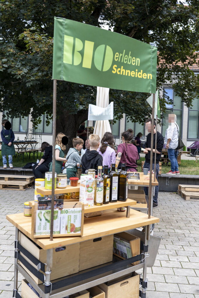 Im Hintergrund steht eine Gruppe von Kindern um einen Wagen herum. Im Vordergrund steht ein weiterer Wagen, mit dem Schild: BIOerleben, Schneiden.