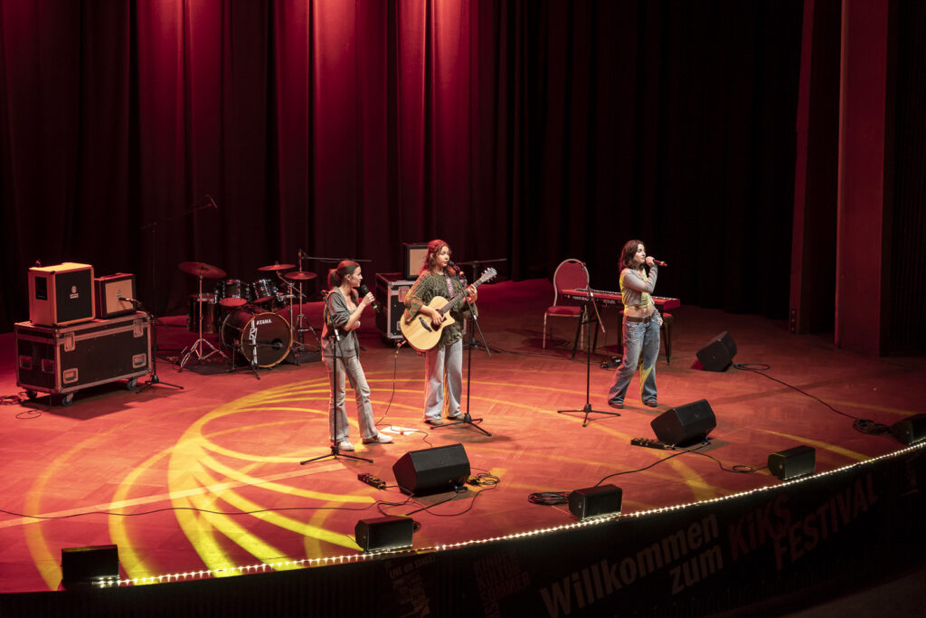 Die drei Mitglieder der Band auf der Bühne, zwei singen, eine spielt Gitarre.