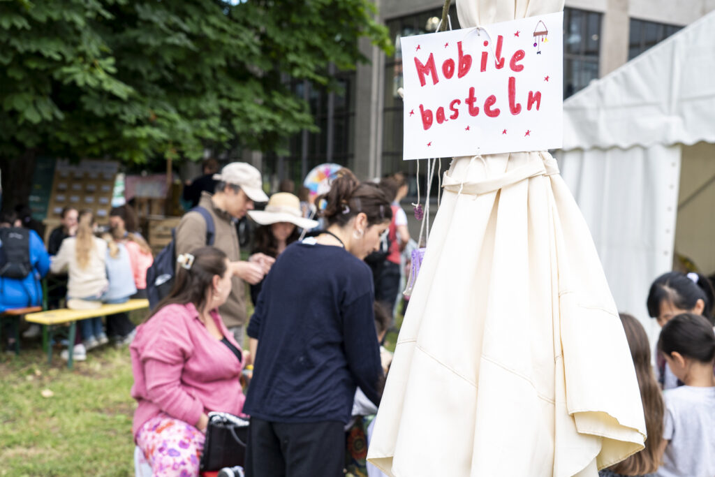 Ein Schild auf dem steht: Mobile basteln. Dahinter sieht man verschwommen ca. 10 Menschen