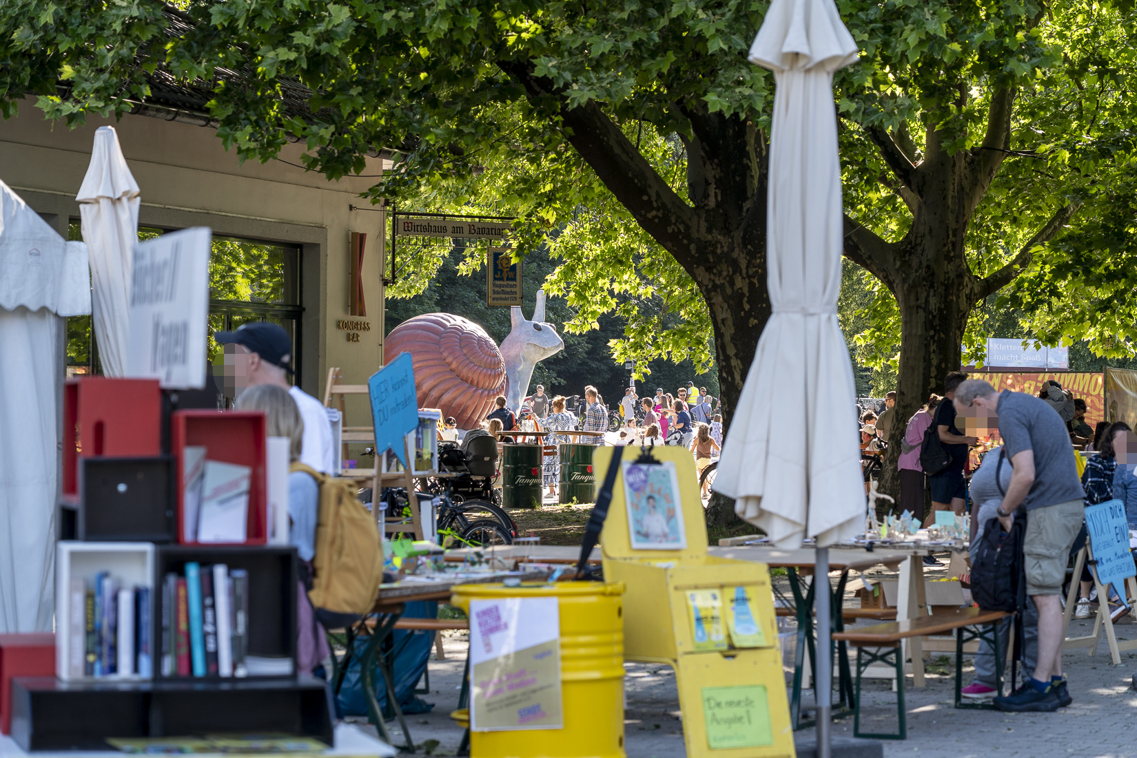 Im Vordergrund mehrere Tisch und ein Zeitungsständer, im Hintergrund viele Menschen in der Sonne auf einem Platz